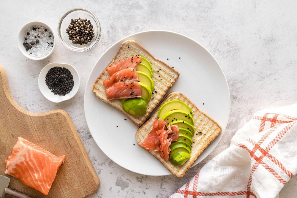 Тарелка с тостами, на которых куски жирной красной рыбы и нарезанный авокадо. Тосты обсыпаны черными кунжутными семечками