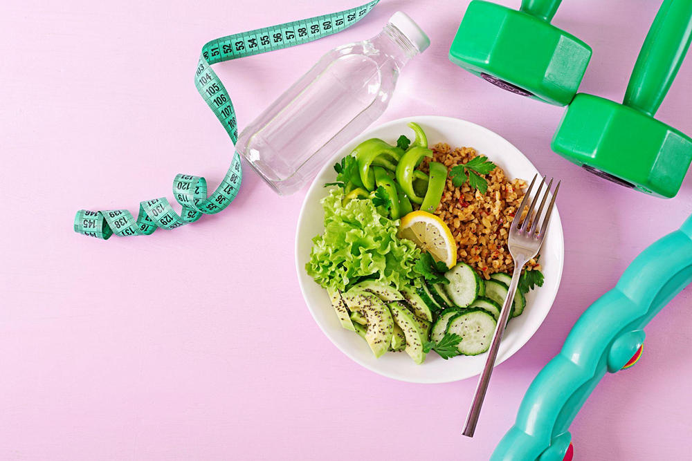 Тарелка с гречкой, нарезанными огурцами, кусочками авокадо, салатом и петрушкой на столе. Рядом бутылка с водой, обруч, гантели и сантиметровая лента
