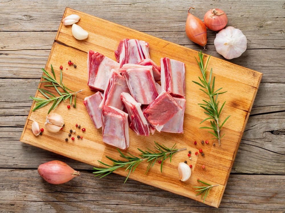 На деревянной доске лежат порезанные кусочки баранины, рядом зелень, лук, чеснок и горошины перца
